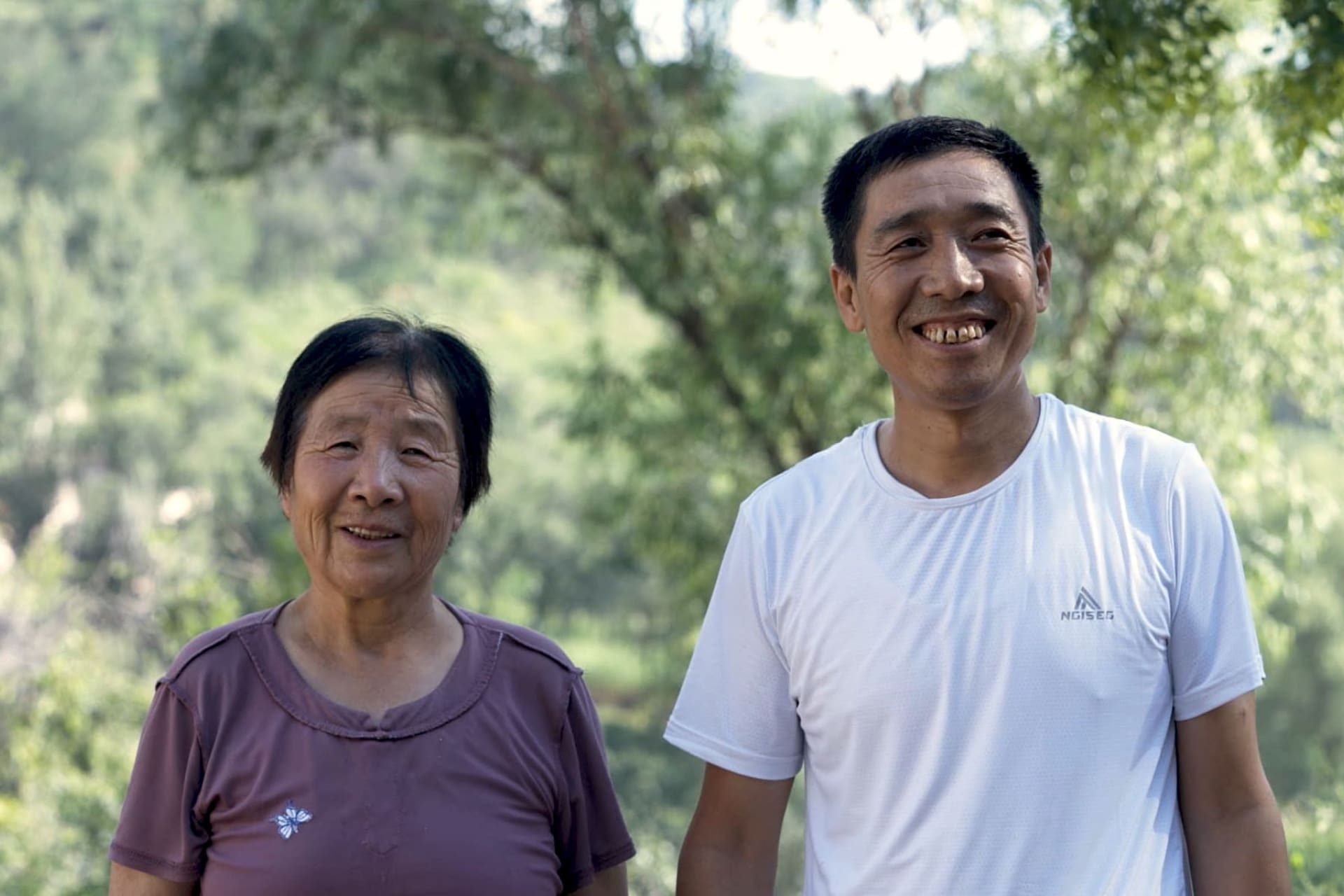 A Chinese woman and Chinese man standing next to each other in front of a green foliage background, smiling at the camera.