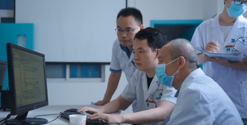 Three male doctors sitting together looking at data on a computer screen. Another male doctor stands off-center behind them. 