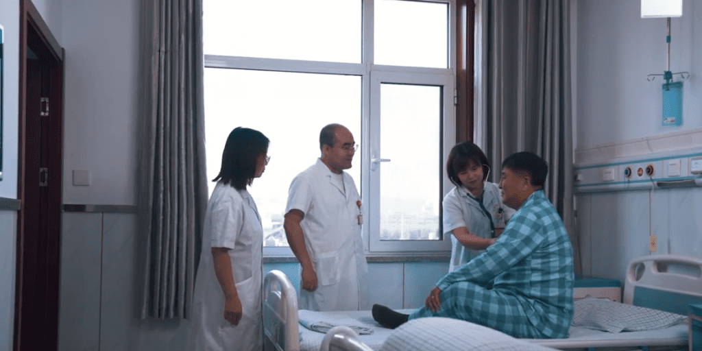 One male doctor and two female doctors standing around a hospital bed speaking with a patient. One doctor is using a stethoscope to listen to the patient's heart. 