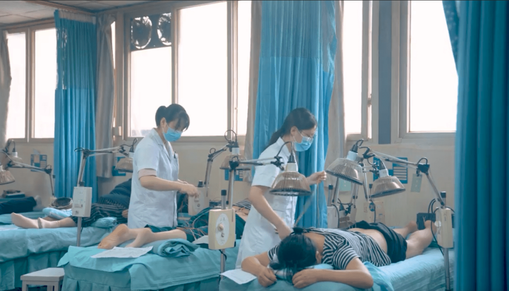 Three patients lie in a row on exam tables in a clinic. Two female doctors stand over two of the patients administering acupuncture. 